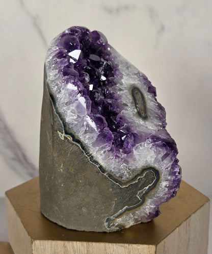 A polished amethyst geode slice with deep purple crystals and a gray matrix, mounted on a light wood block against a white marble background.