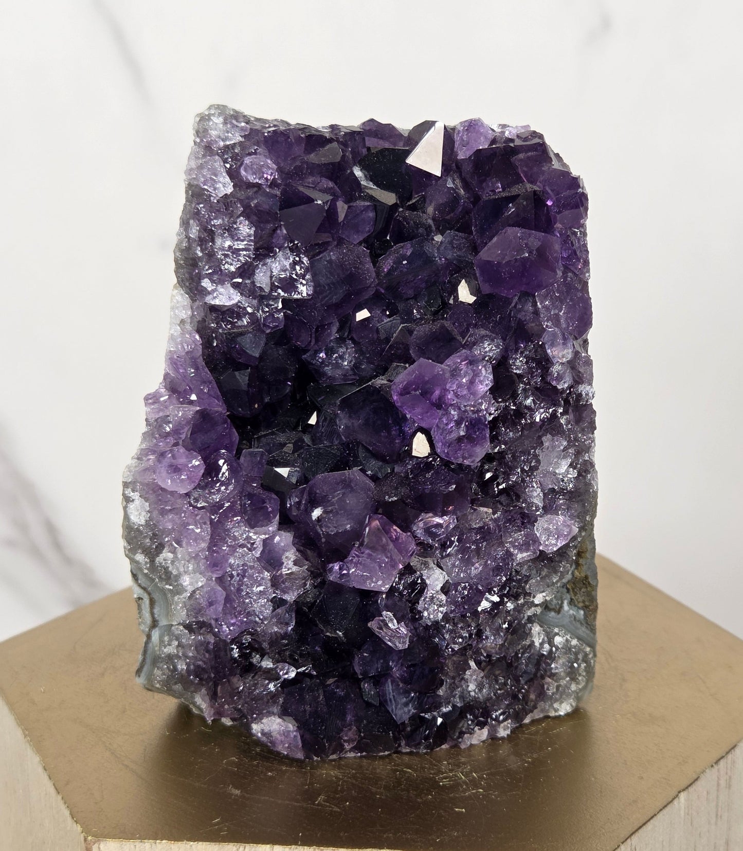 A raw amethyst geode cluster with deep purple and light lavender crystals, mounted on a light wood block against a white marble background.