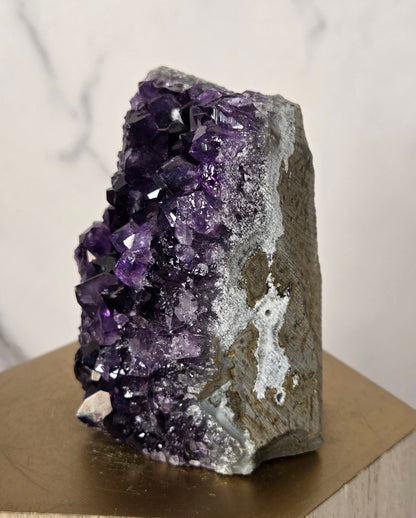 A raw amethyst crystal cluster with deep purple and white crystalline formations, mounted on a light wood geometric display stand against a white marble background.