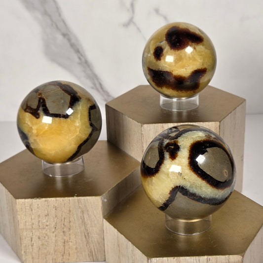 Three decorative septarian spheres  in beige, brown, and black, on a geometric hexagonal wooden wall plate on a white marble-look wall.