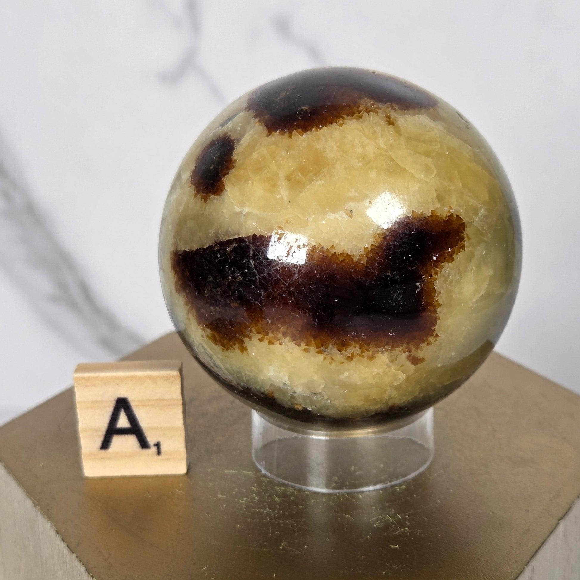 A close-up of a decorative spherical object with a marbled beige, brown, and black pattern, mounted on a light wood geometric stand with a small wooden letter tile labeled 'A' in front.