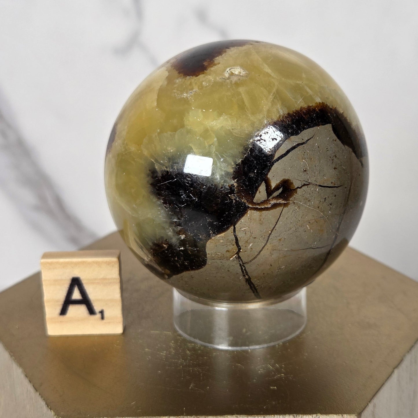 A close-up of a polished stone sphere with a dark inlay, mounted on a light wood block with a small wooden letter tile marked 'A' in front, against a white marble background.