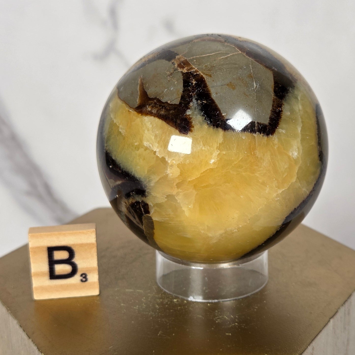 A close-up of a decorative spherical crystal or stone ornament with a marbled yellow, brown, and black pattern inside a clear glass or acrylic sphere, mounted on a light wood block. A small wooden Scrabble tile with the letter B is placed near the base against a light marble-like background.