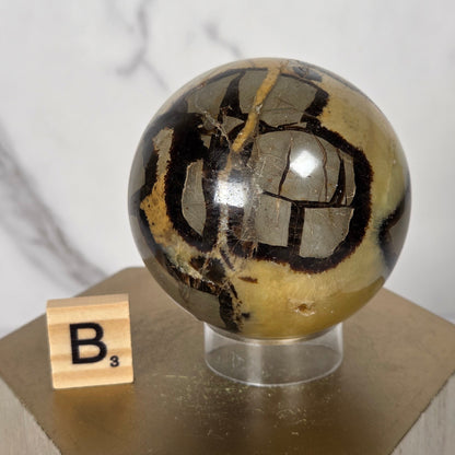 A polished metallic gold and black patterned sphere mounted on a light wood block, with a small wooden tile marked 'B' resting beside it against a white marble-patterned background.