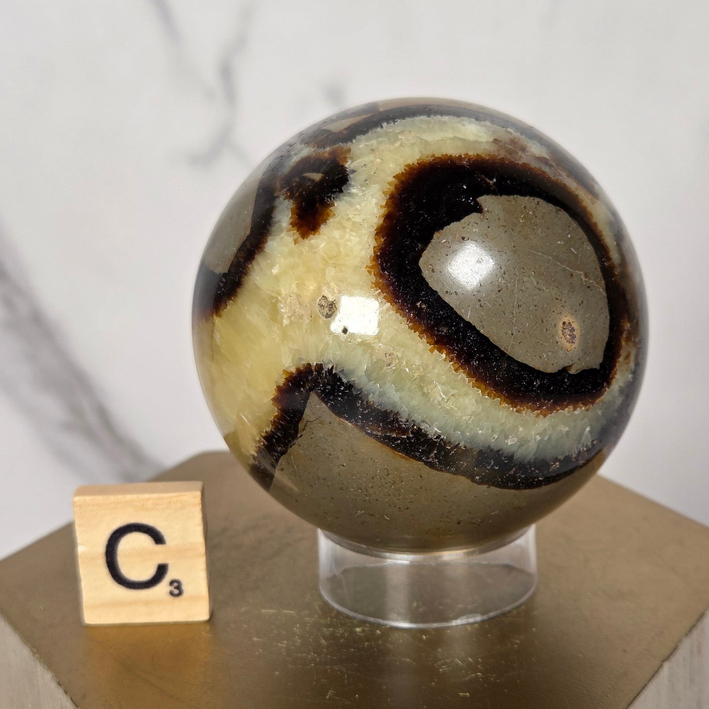A polished stone sphere with swirling black, cream, and brown bands mounted on a clear stand attached to a light wood block, with a small tile showing the letter C against a white marble-like background.