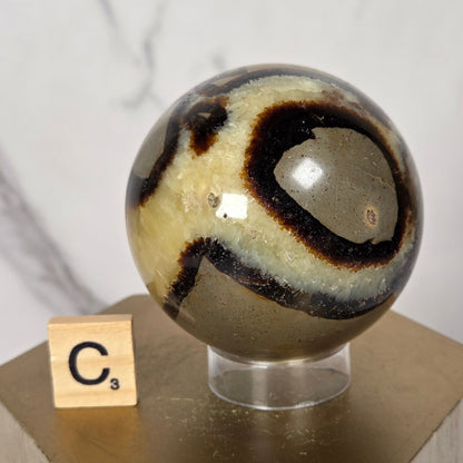 A polished stone sphere with swirling black, cream, and brown bands mounted on a clear stand attached to a light wood block, with a small tile showing the letter C against a white marble-like background.