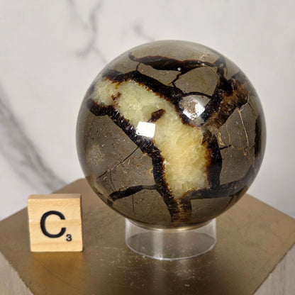 A close-up of a decorative crystal sphere with a metallic gold and cream coloration, mounted on a hexagonal wooden block with a small Scrabble tile showing the letter C.