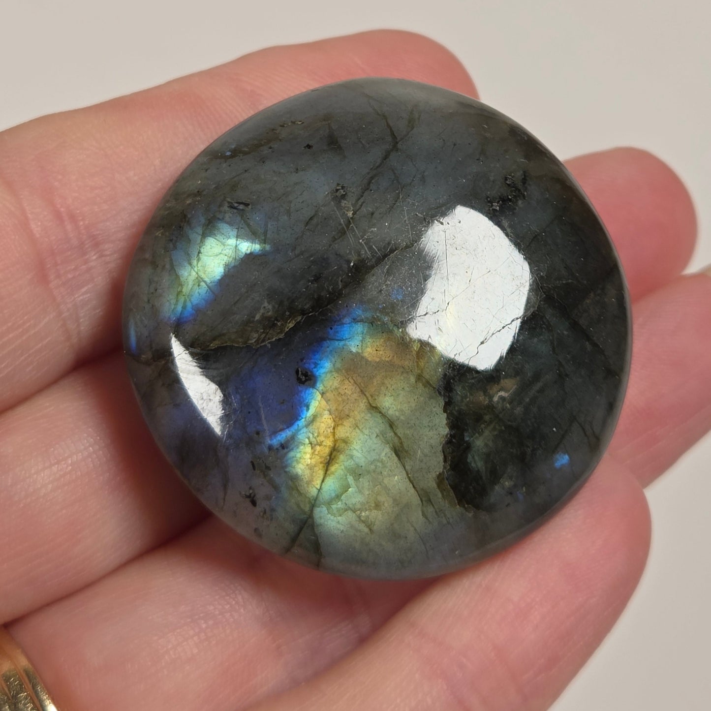 A polished round labradorite gemstone held in a person's hand, showing a gray base with iridescent blue, green, and gold flashes. The person is wearing a gold ring on their right hand.