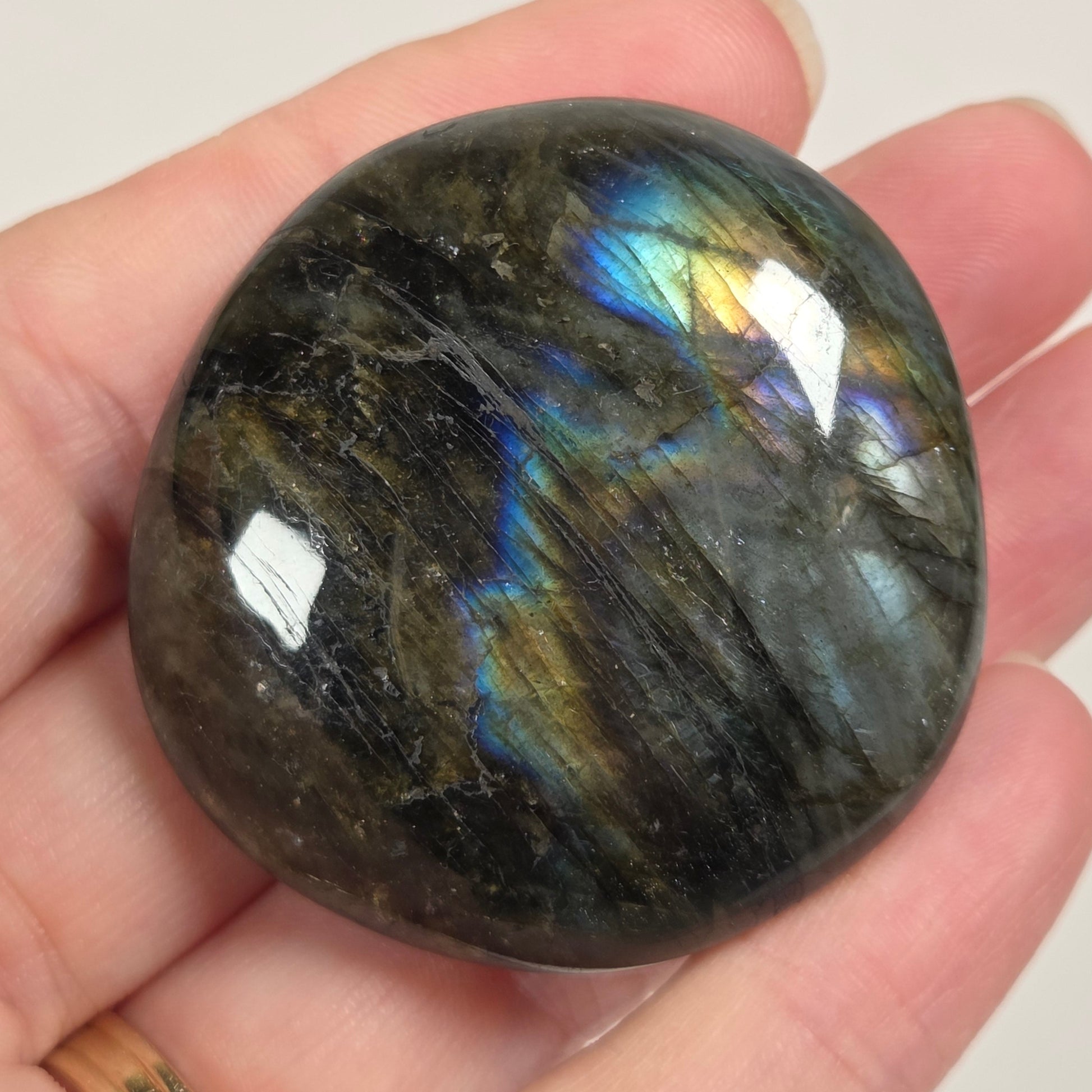 A polished labradorite gemstone with iridescent blue, green, and gold flashes, held in a person's hand against a plain light background.