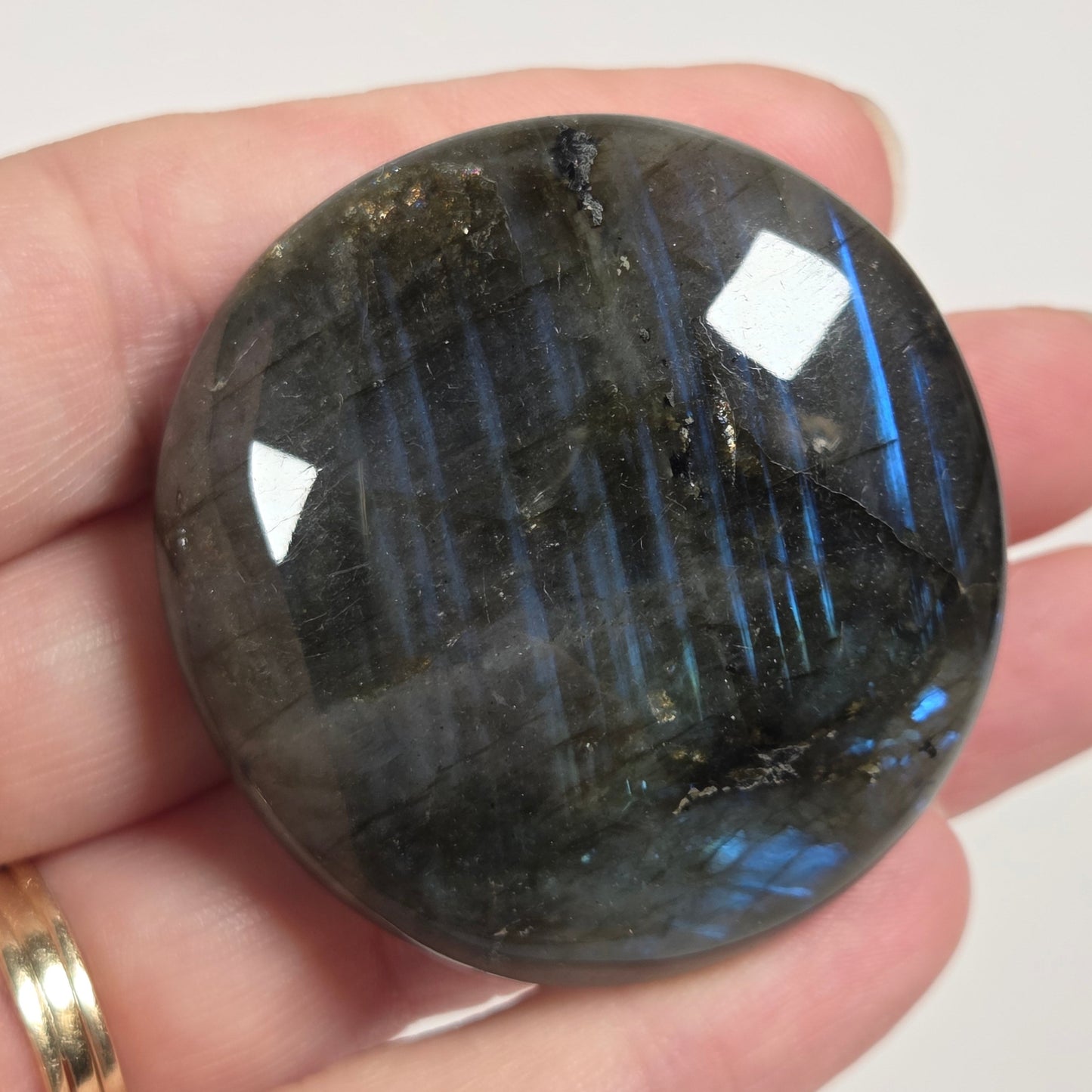 A polished dark gray labradorite gemstone held in a person's hand, showing characteristic blue flashes and white specular highlights.