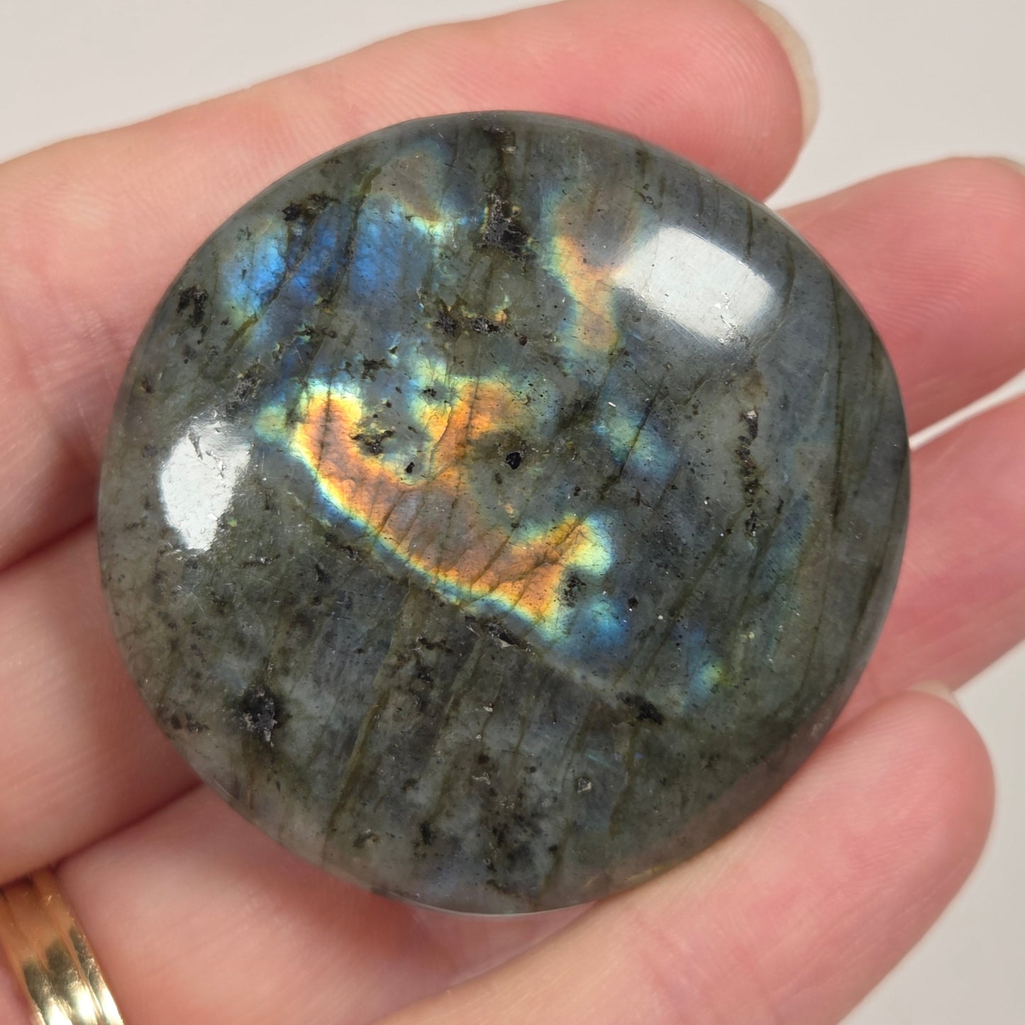 A polished round labradorite gemstone held in a person's hand, showing a gray base with iridescent flashes of blue, green, and gold. The person is wearing a gold band ring on their finger.