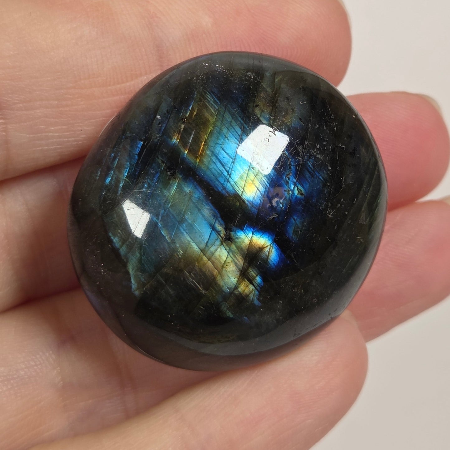 A polished round labradorite stone held in a person's hand, showing a dark gray base with vibrant blue, green, and gold iridescent flashes on a plain light background.