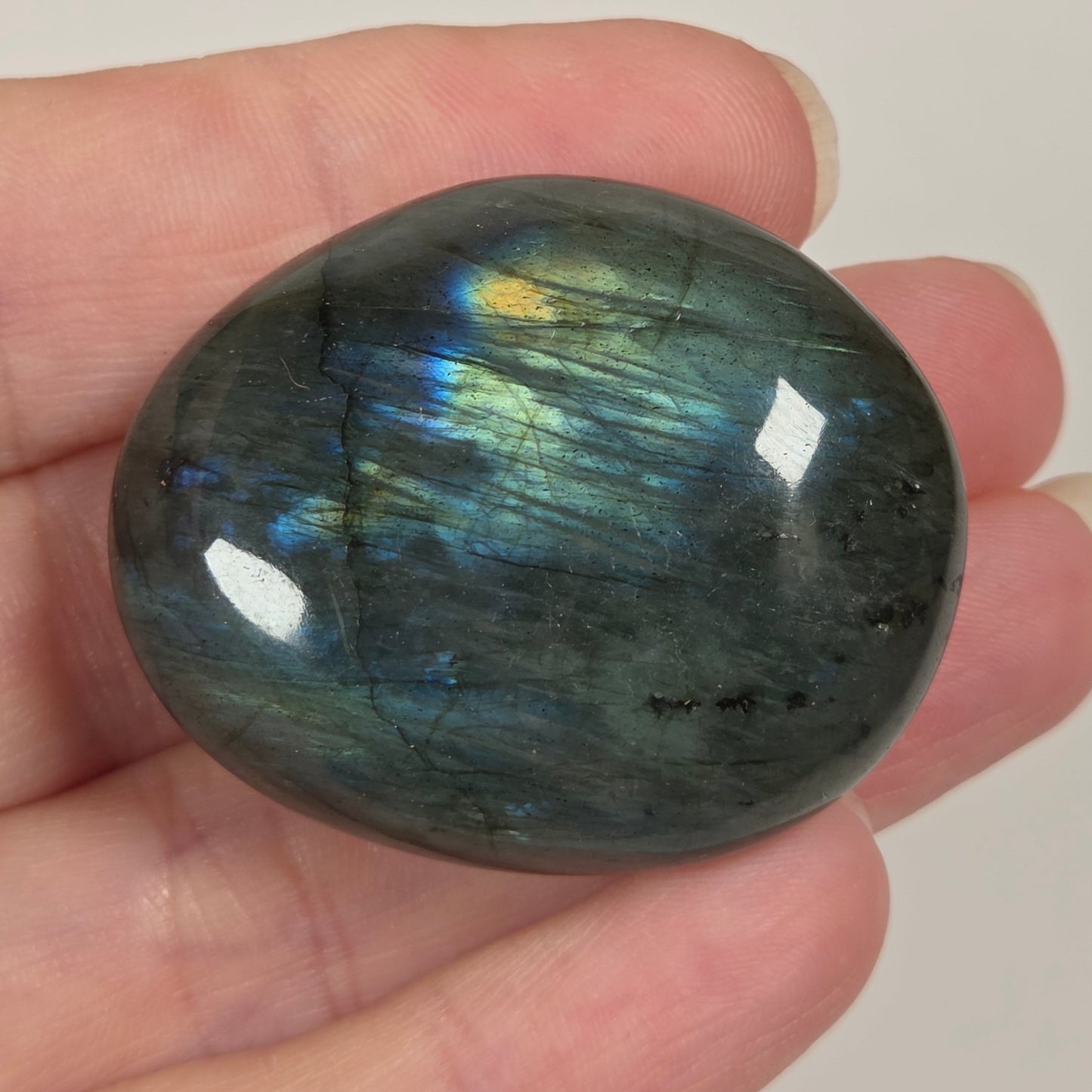 A polished oval labradorite stone held in a person's hand, showing a dark gray base with shimmering blue, green, and gold iridescence.