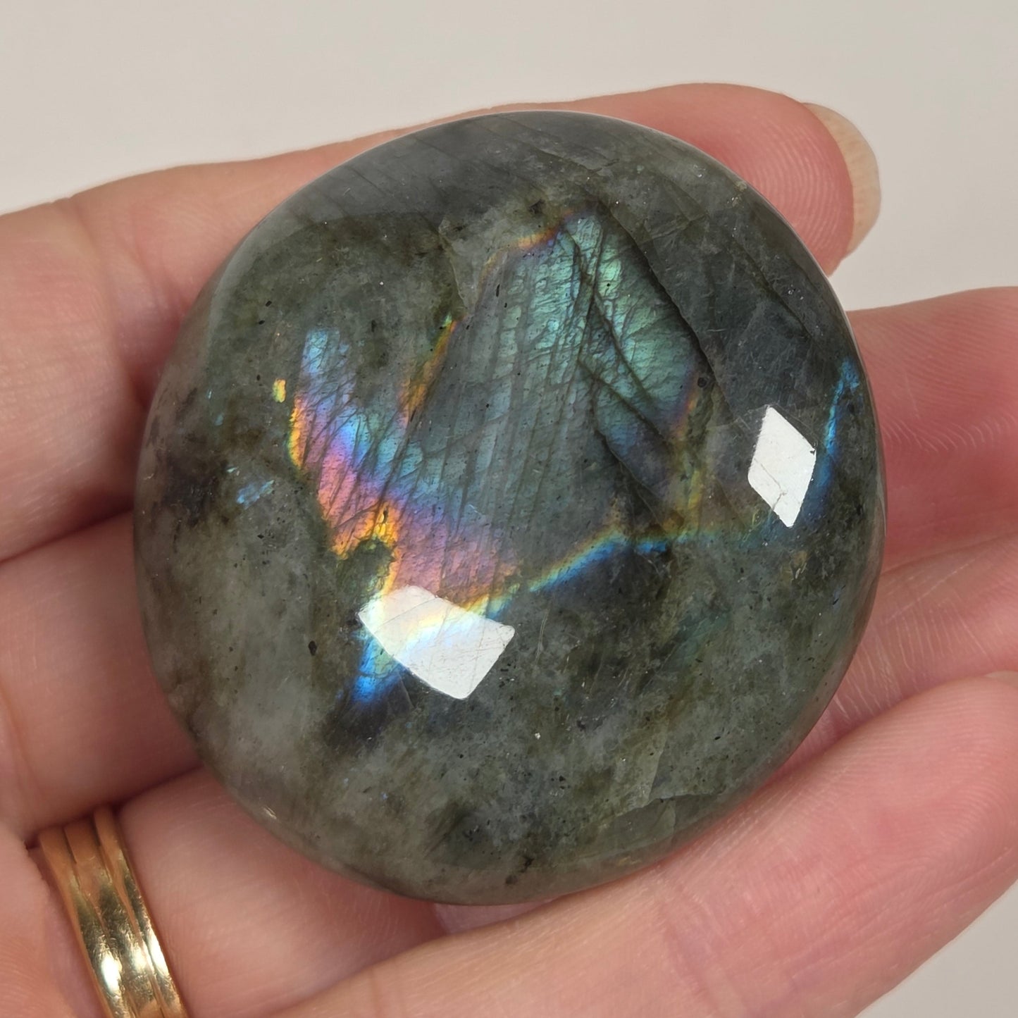 A polished round labradorite stone held in a person's hand, showing a gray base color with vibrant iridescent rainbow flashes and a gold ring on the finger.