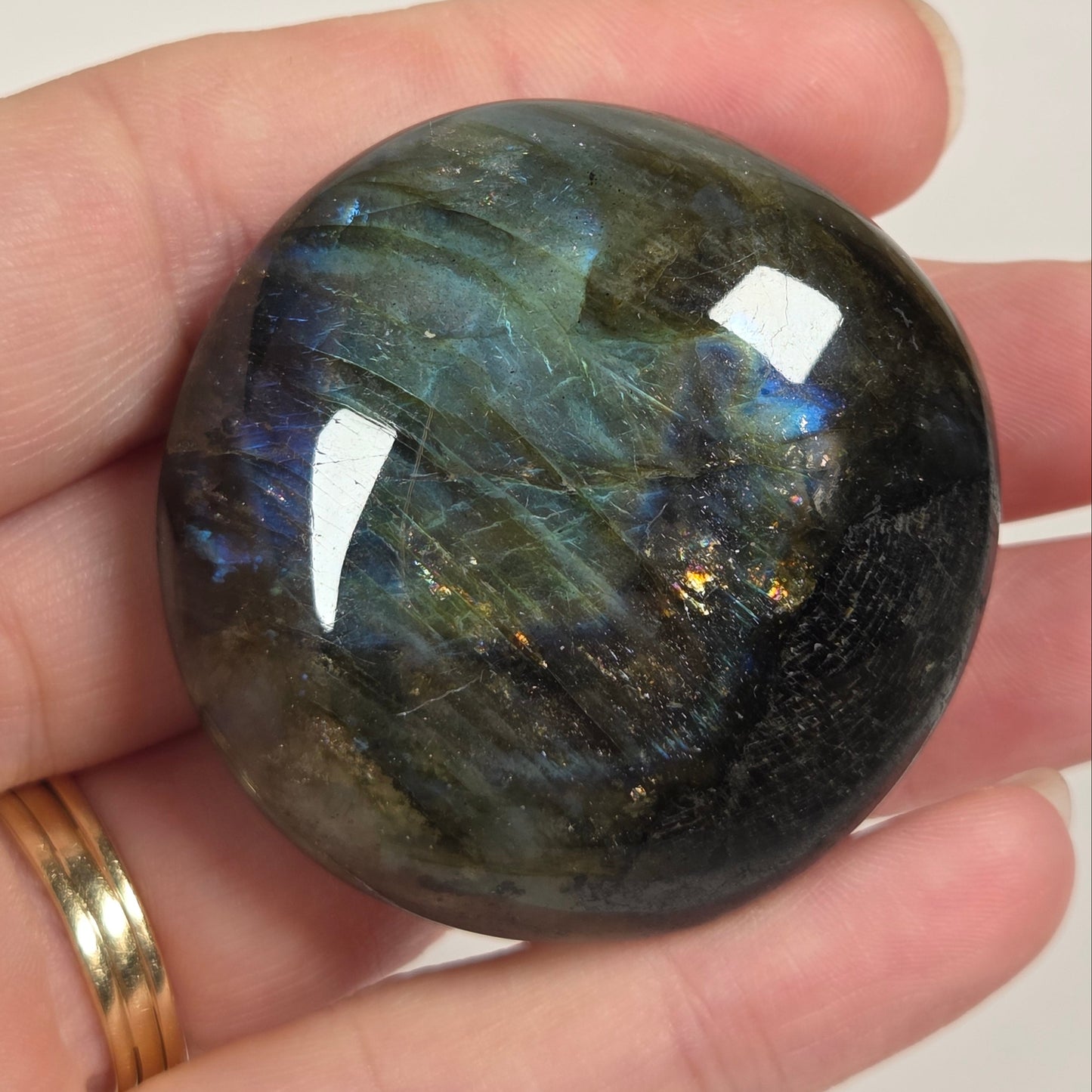 A polished round labradorite crystal ball held in a person's hand, showing a dark gray base with vibrant blue, green, and gold iridescent flashes. The person is wearing a gold ring on their finger against a plain light background.