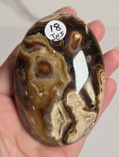 A hand holding a polished oval agate stone with swirling brown, beige, and cream bands, featuring a small white sticker that reads '18 JEZ'.