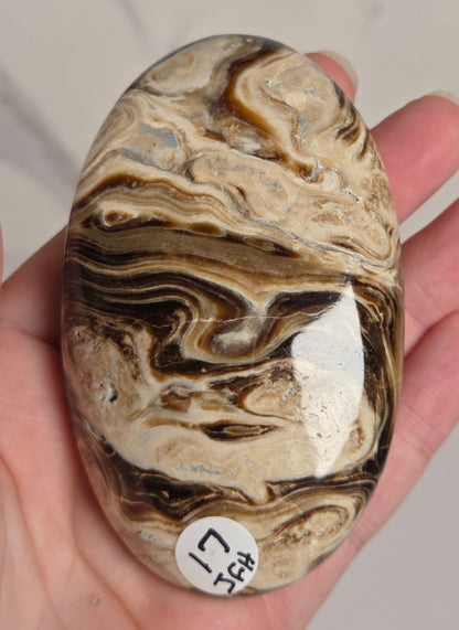 A polished oval stone with marbled swirls of cream, tan, brown, and black, held in a person's hand. A small white sticker with black characters is attached to the right side of the stone.
