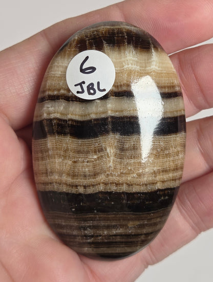 A polished striped stone held in a person's hand. The stone has vertical bands of black, brown, and beige, with a small white circular sticker marked 'JBL 6' attached near the lower left side.