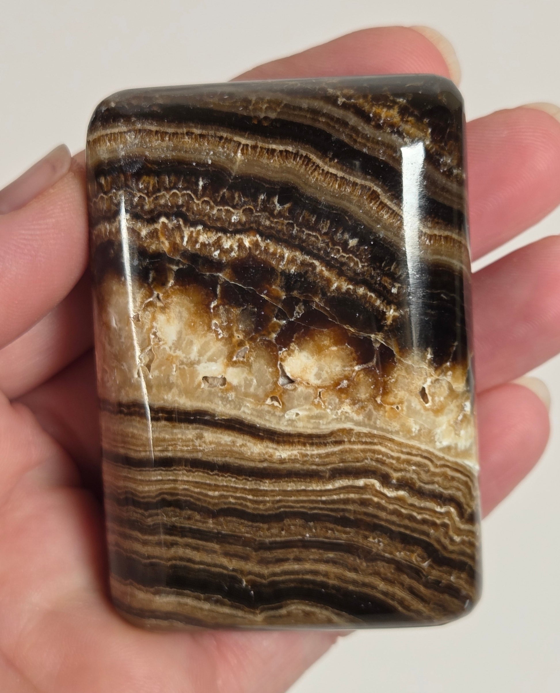 A hand holding a polished rectangular stone with layered brown, black, and cream banding, shown against a plain light background.