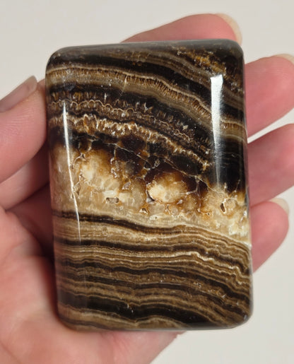 A hand holding a polished rectangular stone with layered brown, black, and cream banding, shown against a plain light background.