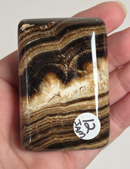 A polished rectangular stone slab with swirling brown, black, tan, and cream banded patterns, held in a person's hand. A small white circular sticker with black handwritten text is attached near the lower right edge.
