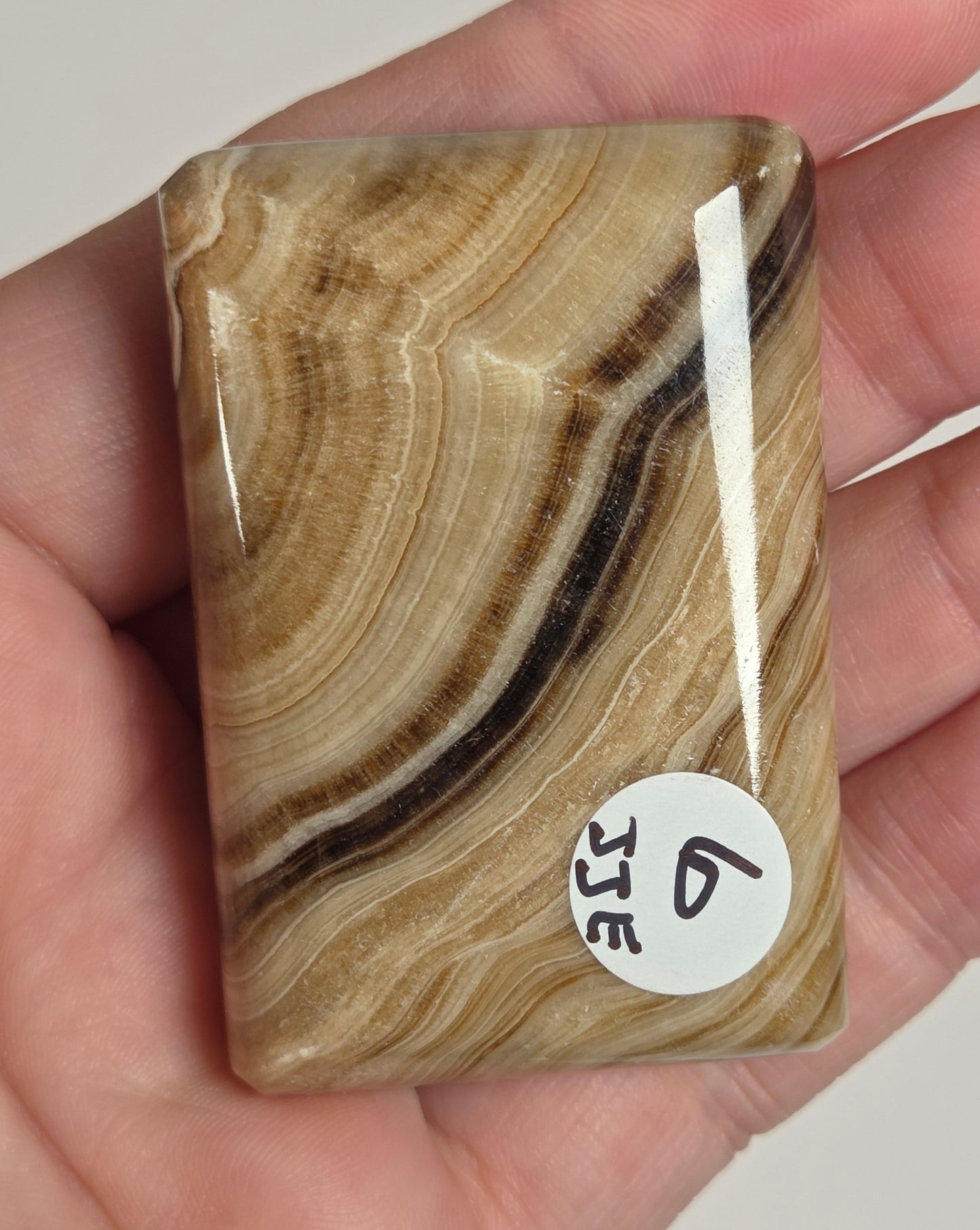 A hand holding a polished rectangular stone slab with natural tan, brown, and cream banded patterns, featuring a small white sticker with the number 6.