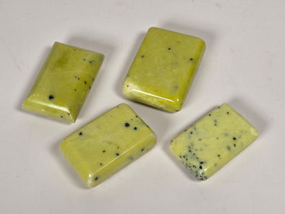 Four rectangular polished stone cabochons with rounded corners, featuring a pale yellow-green color with black speckles, arranged on a plain off-white surface.