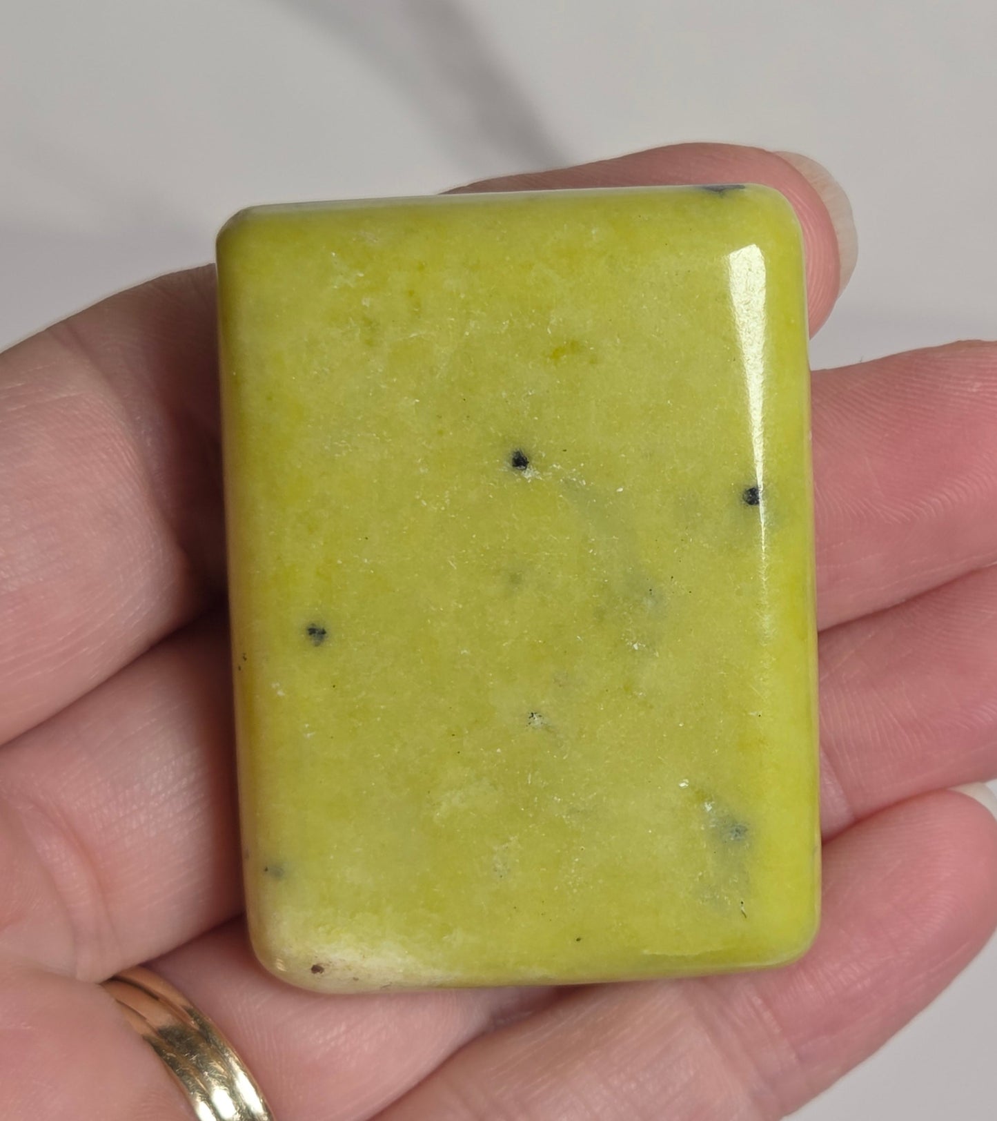 A rectangular yellow-green polished stone or gemstone held in a person's hand, with small dark inclusions visible inside. The hand is shown against a light marble-like background.