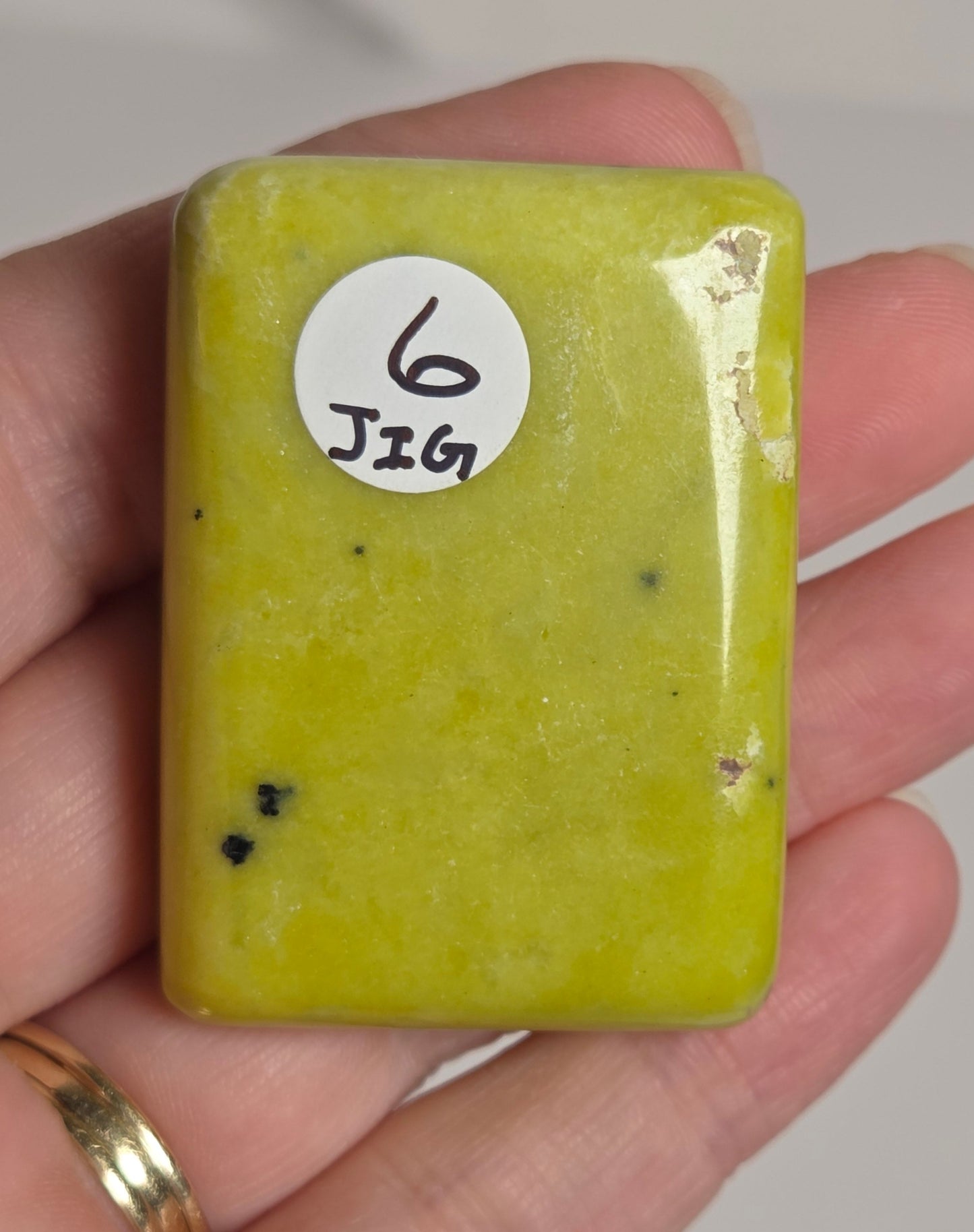 A hand holding a small, rectangular, polished yellow-green stone with a white circular sticker labeled '6 J26'.
