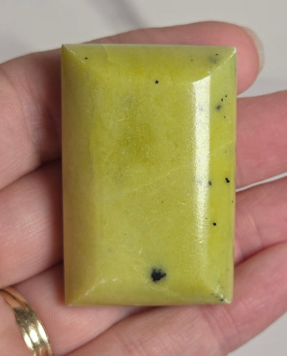 A rectangular pale yellow-green polished stone or gemstone with a few small dark inclusions, held in a person's hand against a light neutral background.