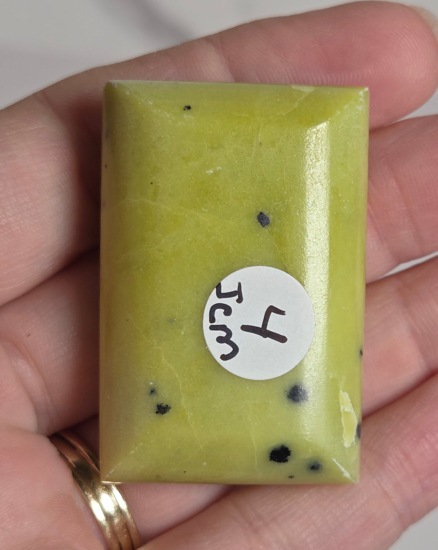 A hand holding a small rectangular bar of soap in a yellow-green color with small black specks. A white circular sticker on the front reads '4 Jcm'.