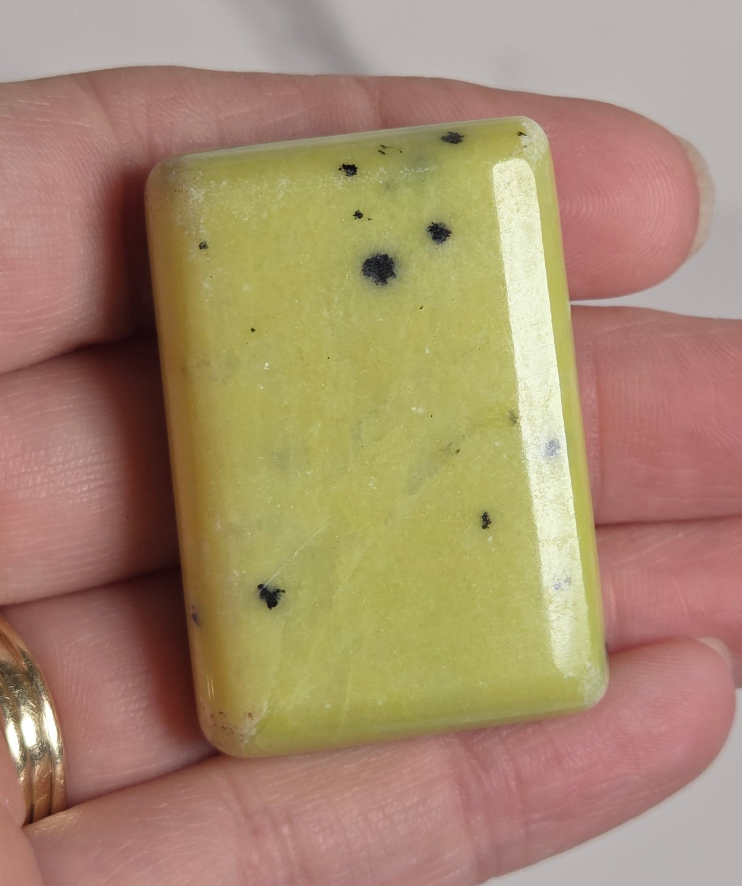 A rectangular pale yellow polished stone or crystal with a few small black specks, held in a person's hand against a light neutral background.