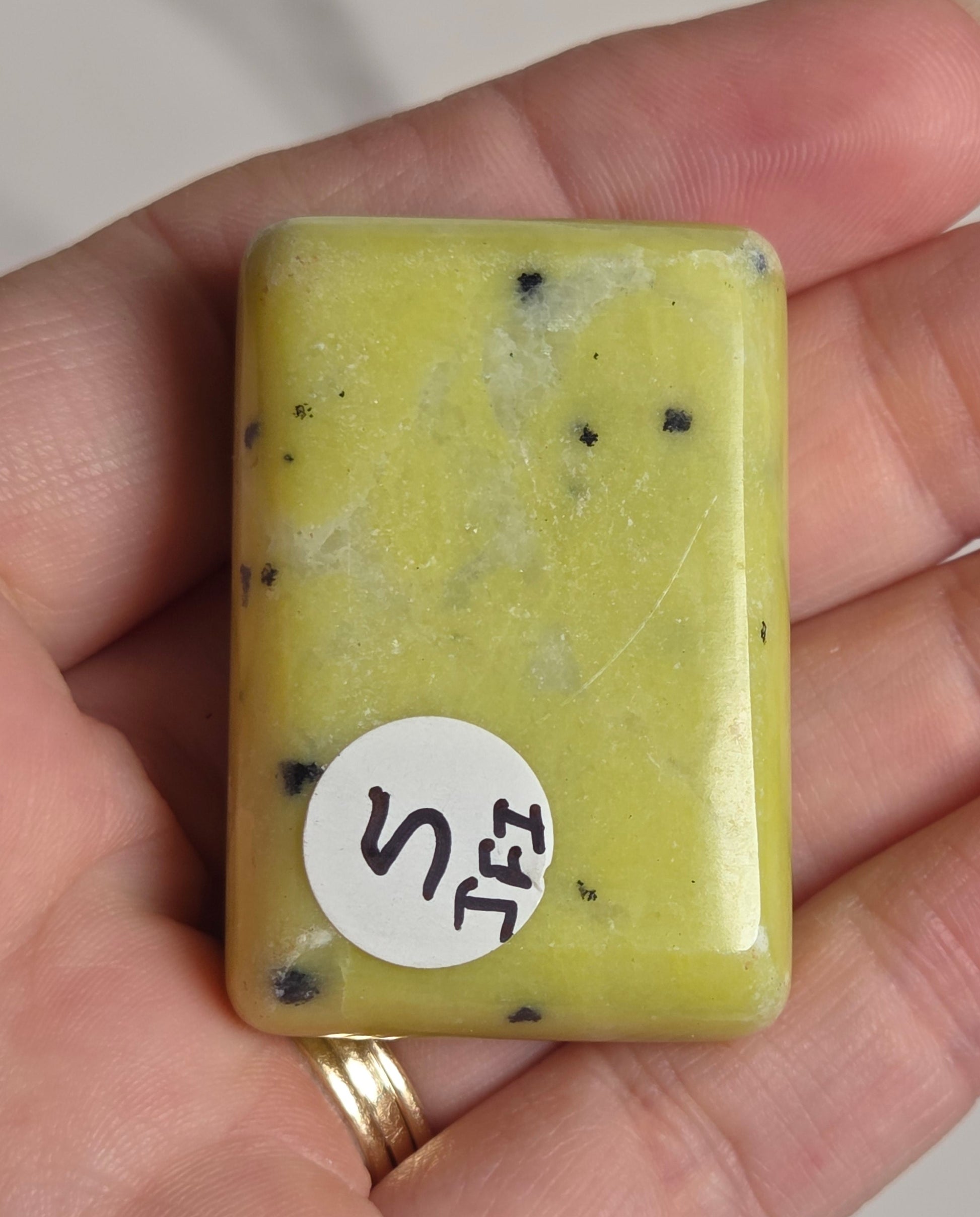 A hand holding a rectangular yellow stone or resin bar with small black speckles and a white circular sticker labeled 'S'.