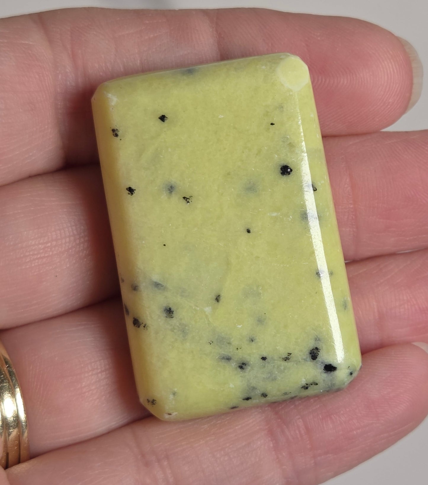 A rectangular pale yellow polished stone or soap slab with scattered small black specks, held in a person's hand against a light neutral background.