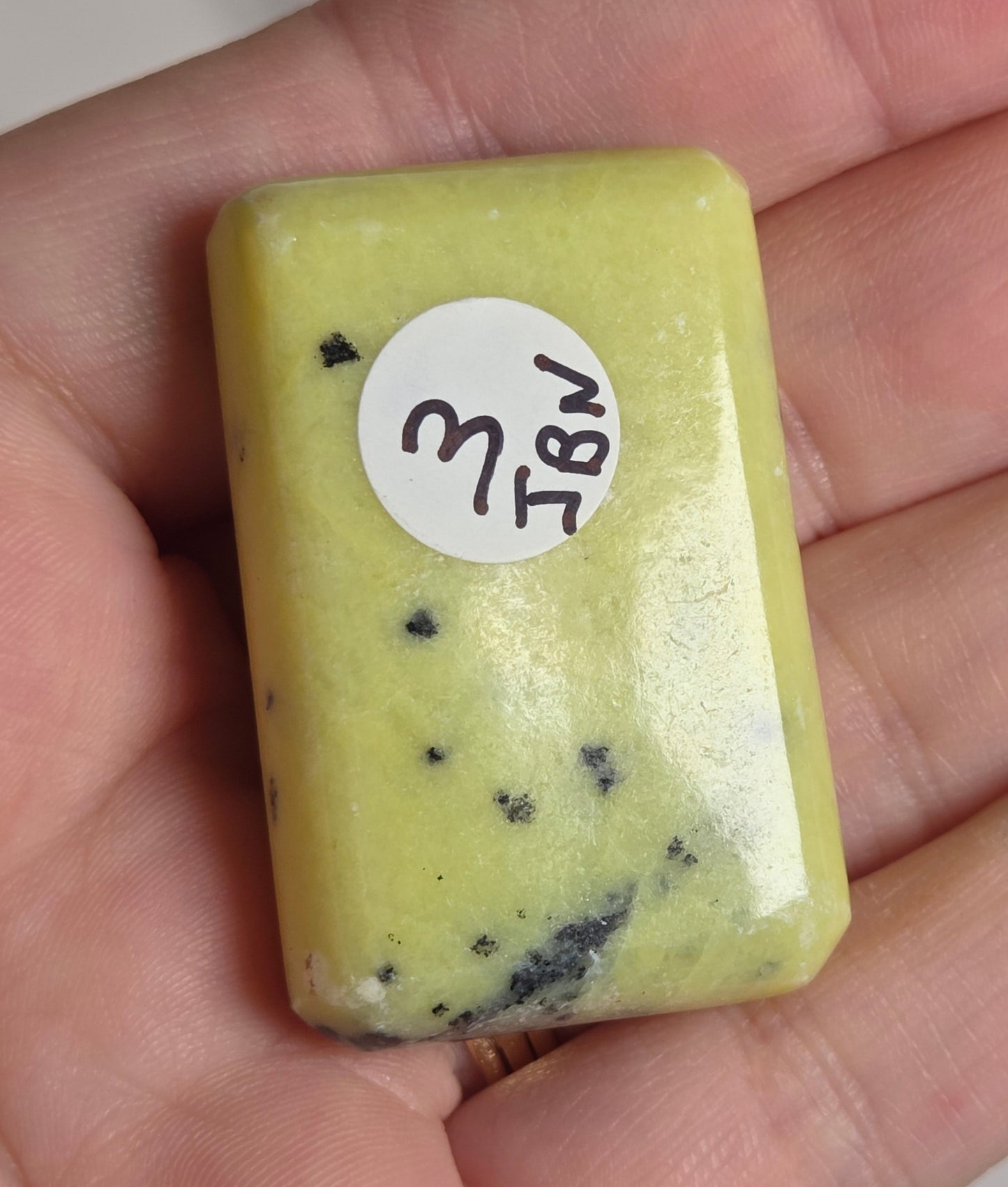 A hand holding a small rectangular pale yellow polished stone or slab with a white circular handwritten label.