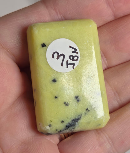 A hand holding a small rectangular pale yellow polished stone or slab with a white circular handwritten label.