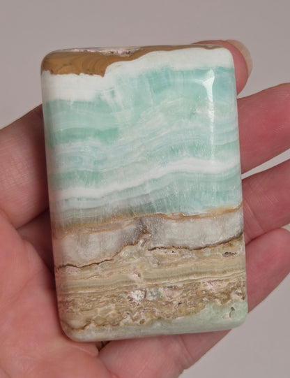 Banded calcite crystal carved into the shape of a soap bar with blue, green and brown coloring being held in a hand on a white background