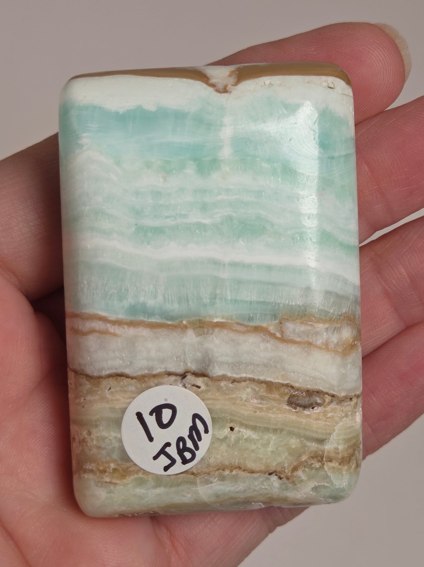 Banded calcite crystal carved into the shape of a soap bar with blue, green and brown coloring being held in a hand on a white background