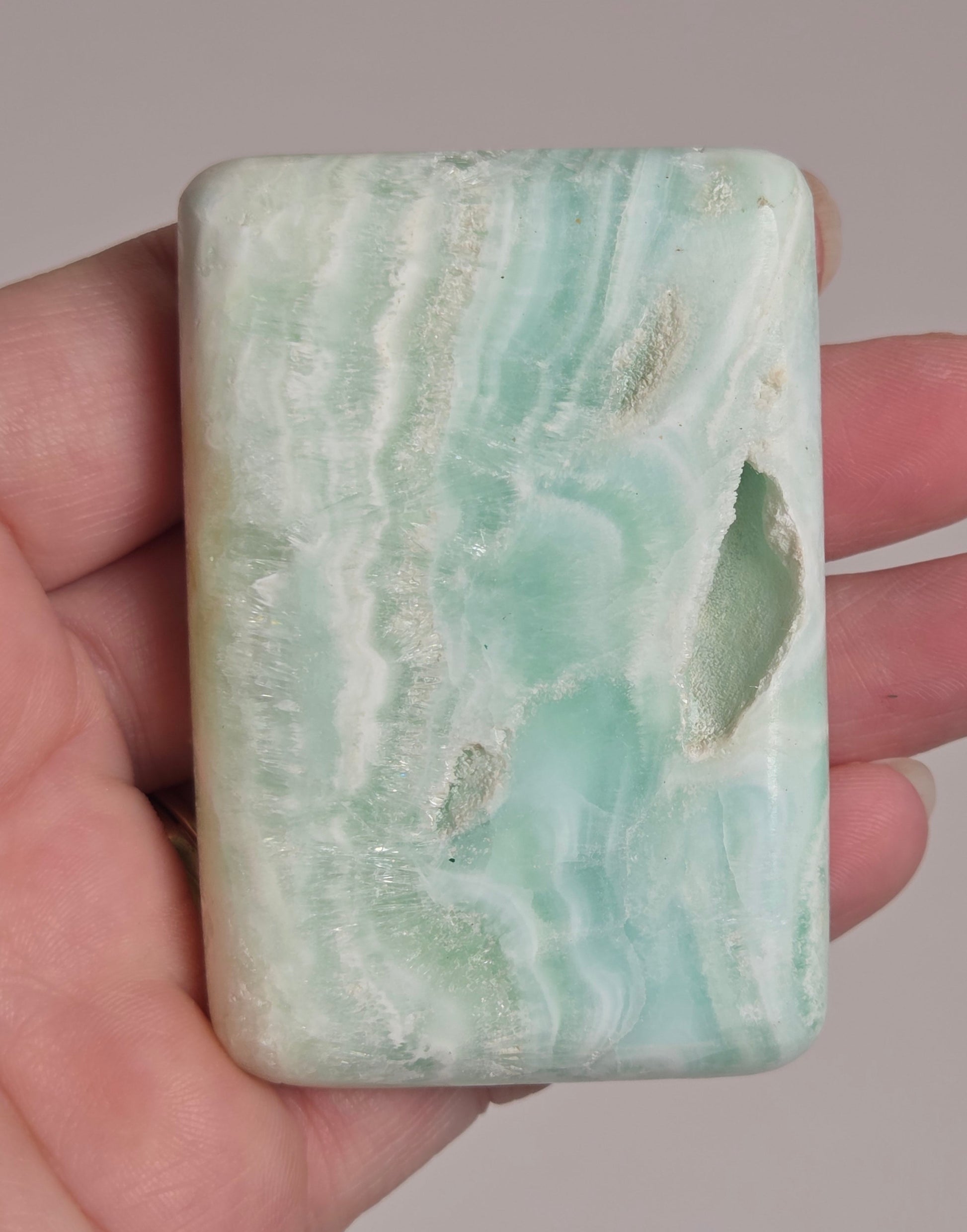 Banded calcite crystal carved into the shape of a soap bar with blue, green and brown coloring being held in a hand on a white background