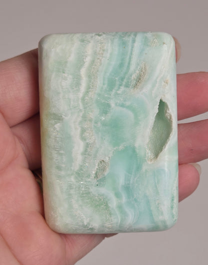 Banded calcite crystal carved into the shape of a soap bar with blue, green and brown coloring being held in a hand on a white background
