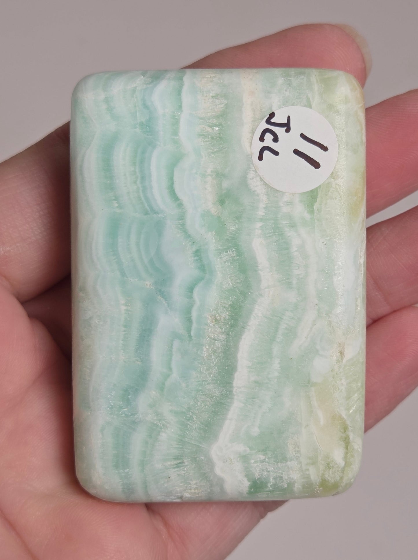 A hand holding a rectangular polished stone slab with wavy light green, white, and pale blue banding, featuring a small white sticker that reads '11 JCL'.