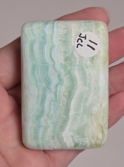 A hand holding a rectangular polished stone slab with wavy light green, white, and pale blue banding, featuring a small white sticker that reads '11 JCL'.