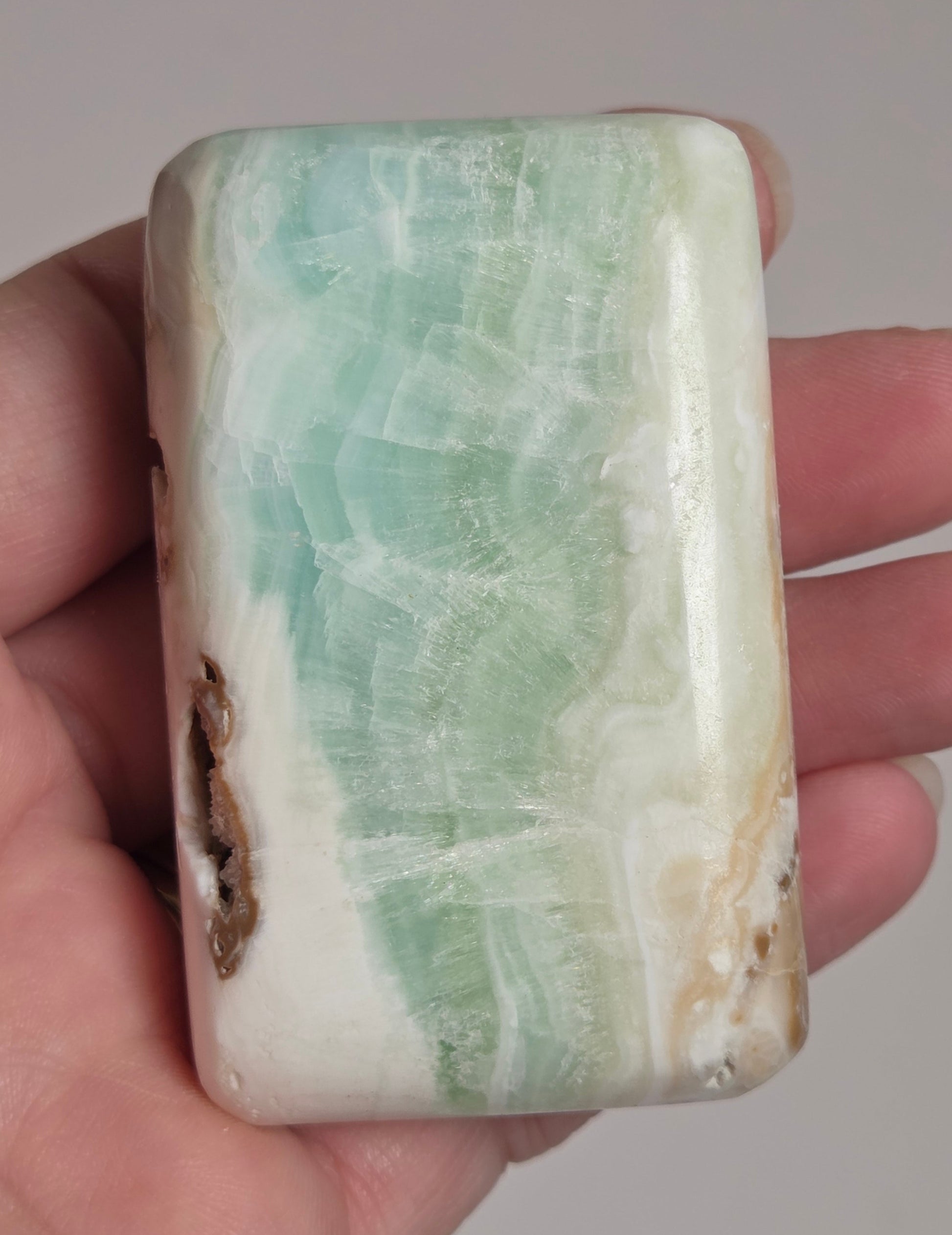 Banded calcite crystal carved into the shape of a soap bar with blue, green and brown coloring being held in a hand on a white background