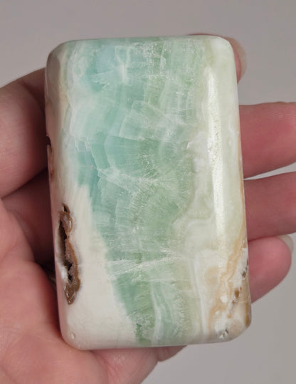 Banded calcite crystal carved into the shape of a soap bar with blue, green and brown coloring being held in a hand on a white background