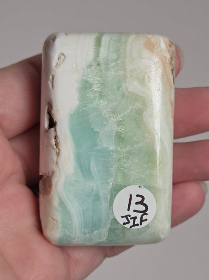 A hand holding a polished rectangular stone with natural marbled bands of pale green, white, and beige, featuring a small white sticker with handwritten numbers on the top right.