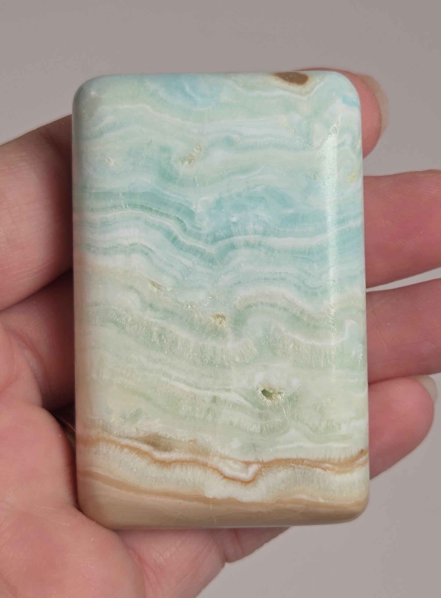 Banded calcite crystal carved into the shape of a soap bar with blue, green and brown coloring being held in a hand on a white background