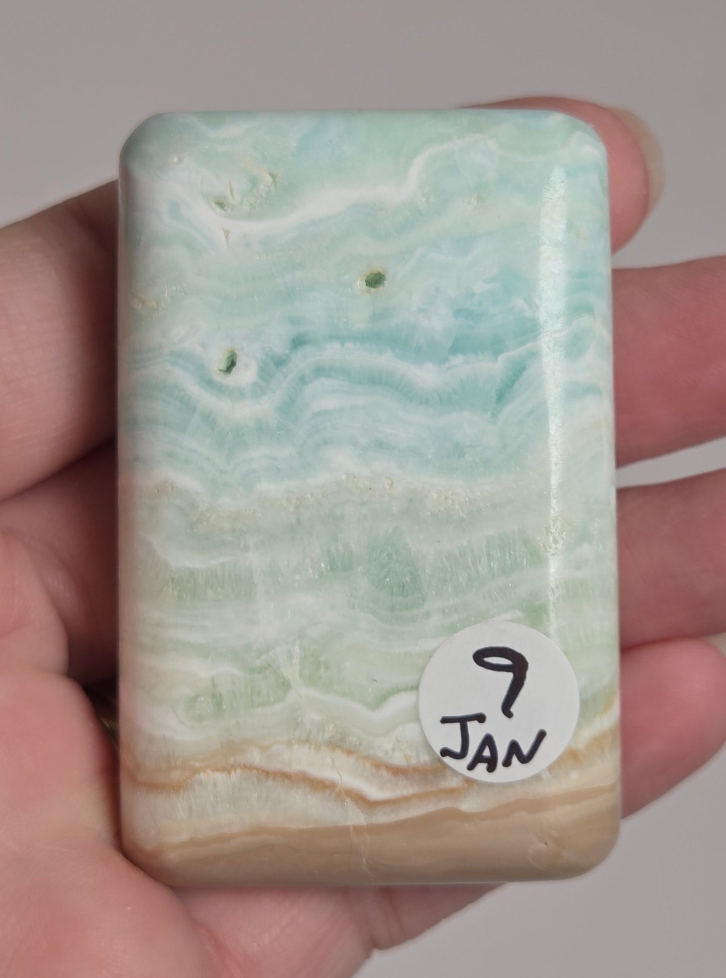 Banded calcite crystal carved into the shape of a soap bar with blue, green and brown coloring being held in a hand on a white background