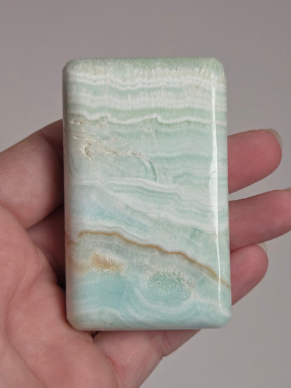 Banded calcite crystal carved into the shape of a soap bar with blue, green and brown coloring being held in a hand on a white background