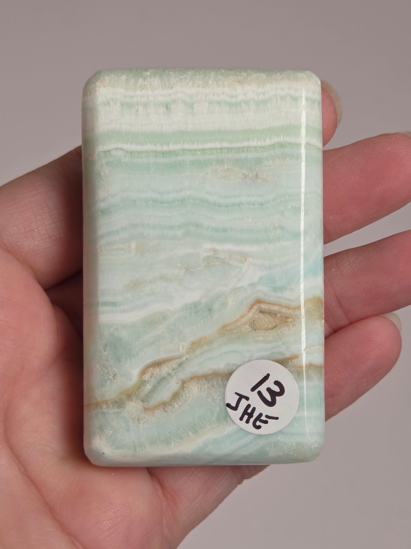 A hand holding a polished rectangular stone tile with pale mint green, white, and beige veining, featuring a small white sticker with handwritten text on the top right.