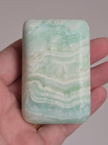 Banded calcite crystal carved into the shape of a soap bar with blue, green and brown coloring being held in a hand on a white background